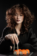 Asian female with curly hair enjoying sushi in elegant black outfit, Fashion photo Philadelphia...