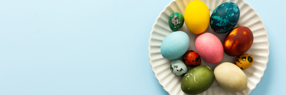 A variety of decorated Easter eggs in vibrant colors are creatively arranged on a white plate set against a soft blue background, symbolizing spring festivities and joy, banner