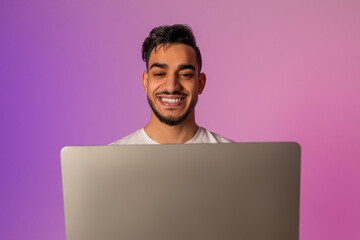 A happy young Arab man is engaged in a video call on his laptop. The colorful neon lights create a lively atmosphere as he enjoys chatting online, connecting with others remotely.