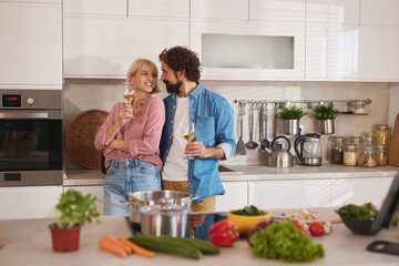 A joyful couple stands close in a modern kitchen, sharing smiles while holding glasses of drink. Fresh vegetables and pots are spread across the counter, creating a lively cooking atmosphere.