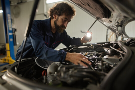 A skilled mechanic is working on a car's engine, using a flashlight to check various parts under the hood. The repair shop is well-equipped and organized, highlighting professional car service.