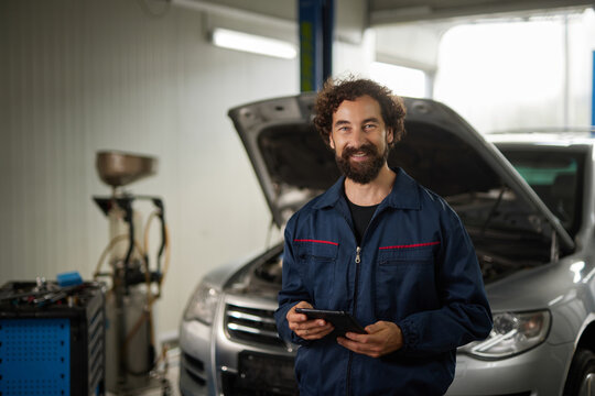 A mechanic with a curly beard stands in a workshop, holding a tablet while working on a silver car with the hood raised. Tools and equipment are visible in the background.