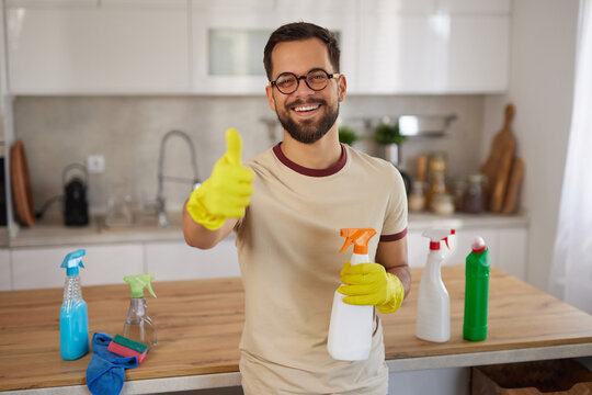 A man wearing yellow gloves smiles and gives a thumbs-up while holding cleaning products in a well-lit kitchen. The space is tidy with vibrant colors from supplies, promoting a clean environment.