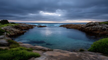 A dramatic cloudy sky looms over a tranquil rocky cove with clear turquoise water at twilight