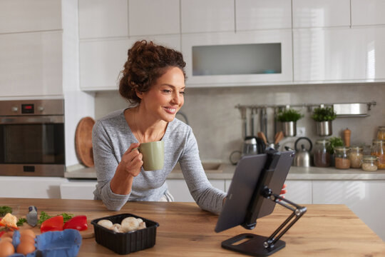 A woman smiles as she holds a cup and looks at a tablet displaying a recipe. Fresh ingredients are neatly arranged on the kitchen counter around her.