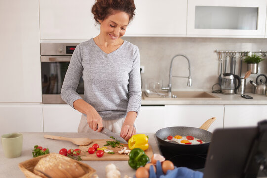 A happy person chops fresh vegetables like bell peppers, tomatoes, and mushrooms on a wooden board. A tablet displays a recipe beside them in a bright kitchen. - Powered by Adobe