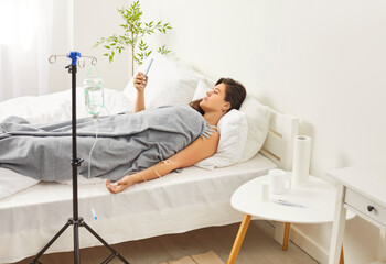 Sick upset young woman lying on bed in bedroom at home receiving IV drip infusion in blood and...