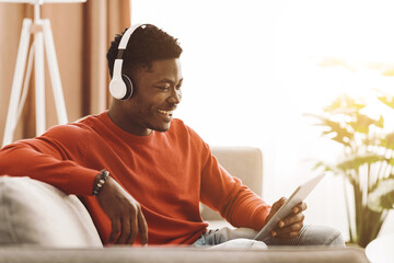 A young man in a cozy living room listens to music through headphones while using a tablet. Sunlight streams in, creating a warm atmosphere and a sense of relaxation. © Prostock-studio
