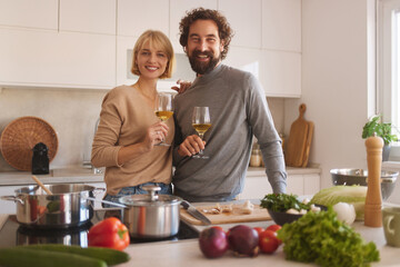 A couple stands in a bright kitchen, smiling and holding wine glasses. Fresh vegetables and cooking tools are on the counter, creating a cheerful cooking atmosphere.