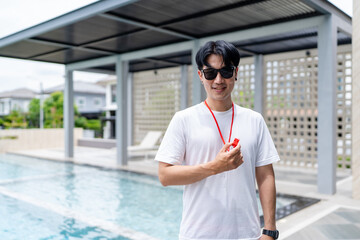 Portrait of asian lifeguard man at swimming pool with whistle ready to save children or seniors from accident, handsome instructor or trainer for swimming sport at clubhouse