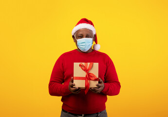 A senior black man wearing a Santa hat and protective mask stands against an orange background, holding a gift box. He looks directly at the camera, embodying holiday spirit during lockdown.