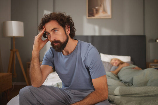 A man sits on the edge of a bed, looking distressed and lost in thought. A woman sleeps peacefully under the covers behind him, creating a contrast of emotions in a quiet bedroom.