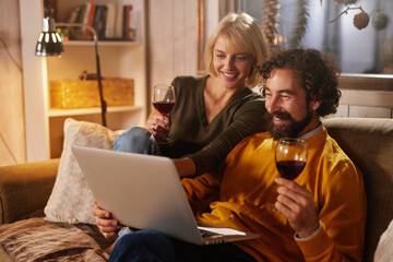 Two people relax on a sofa in a warm-lit room, sharing laughter over a laptop while sipping wine. The cozy setting is perfect for a pleasant evening together.