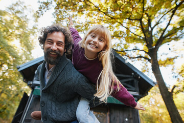 A smiling father gives his daughter a piggyback ride outdoors amidst vibrant autumn leaves. Their joyful expressions capture a special moment together on a weekend.