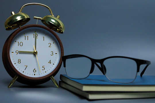 Defocused black glasses resting on a stack of books beside a vintage alarm clock over grey background, simple workspace scene symbolizing study, focus, productivity and time management.