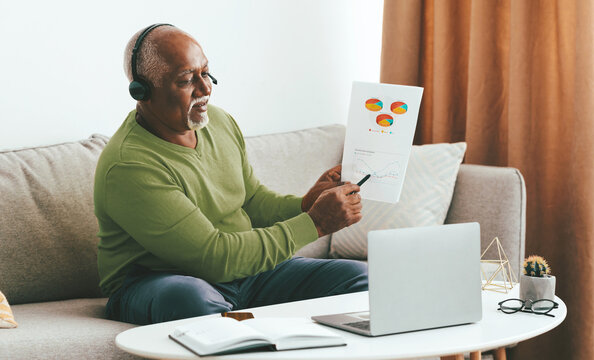 A senior man is engaged in a video call from his living room. He wears a headset and holds a report with colorful graphs. A laptop and open notebook are nearby, indicating his analytical focus.