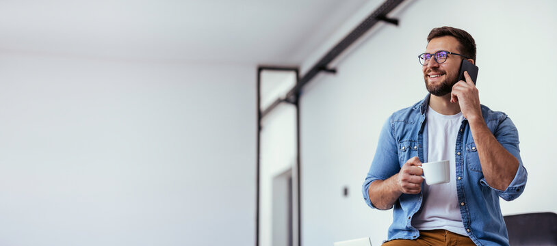 Handsome bearded man leaning on office desk, drinking coffee and talking on the phone. - Powered by Adobe
