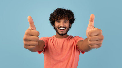 A handsome young eastern guy in a pink t-shirt poses alone against a blue background. He smiles warmly while giving a thumbs up, suggesting he recommends something positive.