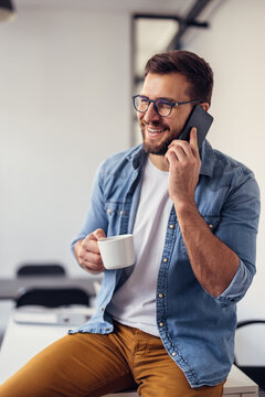 Handsome bearded man leaning on office desk, drinking coffee and talking on the phone.