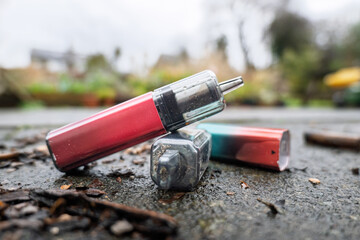 Discarded rechargeable vapes left lying on a wet pavement. One has its battery compartment separated from the head section.