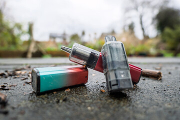 Discarded rechargeable vapes left lying on a wet pavement. One has its battery compartment separated from the head section.