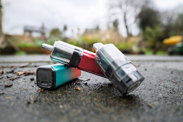 Discarded rechargeable vapes left lying on a wet pavement. One has its battery compartment separated from the head section.