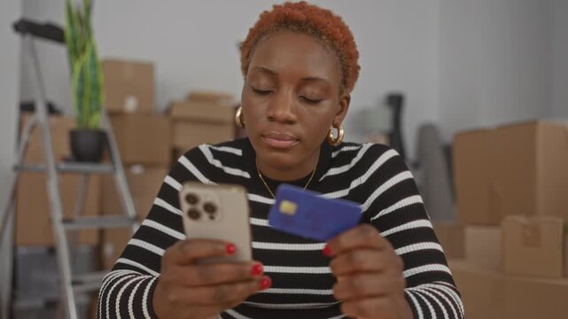 Woman holding creditcard and smartphone amid moving boxes in a building interior; financial planning focus.