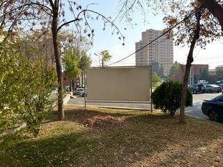 Blank billboard or banner sign standing in an urban park, surrounded by autumn trees and lawn. A multi-story building is visible in the background. Ideal copy space for text or advertising.