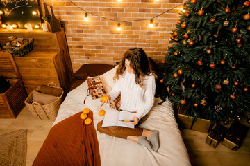 beautiful curly girl in a white sweater with dark hair sits on the bed smiling and writes in a notebook. happy new year portrait, girl makes a wish and writes a letter