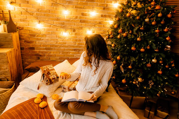 beautiful curly girl in a white sweater with dark hair sits on the bed smiling and writes in a notebook. happy new year portrait, girl makes a wish and writes a letter