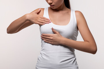 Unrecognizable young woman in a white top examines her breasts in a grey studio background. This closeup emphasizes breast screening awareness and cancer prevention.