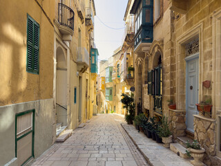 Picturesque view down a classic, gently sloping street in Valletta, Malta. The soft daylight beautifully highlights the detailed limestone facades and the iconic, colorful traditional Maltese wooden b