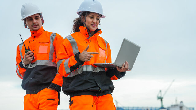 A female and male engineer in orange safety gear and hard hats collaborate at a busy commercial port. The woman holds a laptop while the man uses a radio near a cargo ship. - Powered by Adobe