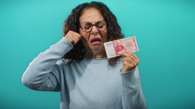 Middle-aged woman in blue sweater on turquoise background holding icelandic krona banknotes with a sad expression reflecting financial distress.