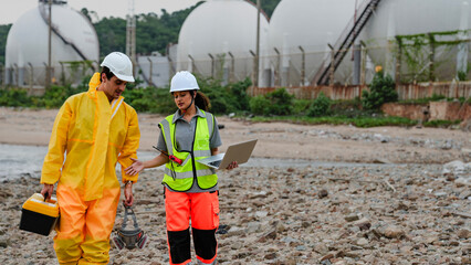 Environmental scientists conduct water quality testing at an industrial discharge point. One in a hazmat suit stands in the water while the other records data on a laptop.