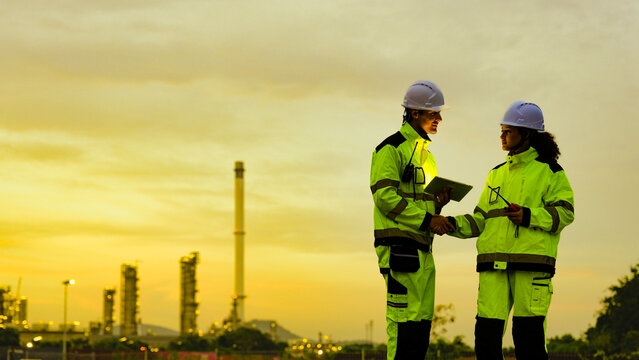 Petrochemical engineers in PPE conduct a site survey at a refinery. They use a tablet for data analysis and operational monitoring during the evening shift.