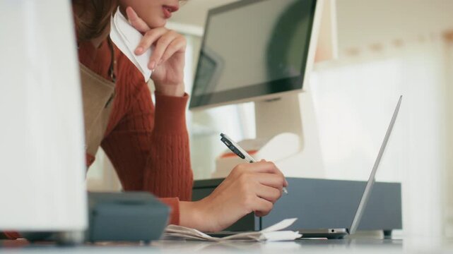 Close up Young Asian woman shop owner writing on notebook, checking bill, calculating budget, income for planning and managing stock inventory in coffee shop. Small business bookkeeping concept.