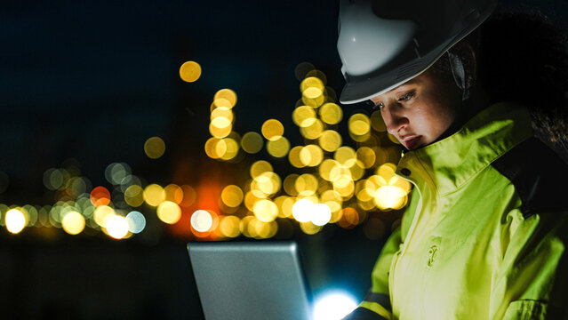 A petrochemical engineer in PPE monitors refinery operations during a night shift. She uses a laptop for real-time data analysis in front of lit-up processing infrastructure. - Powered by Adobe