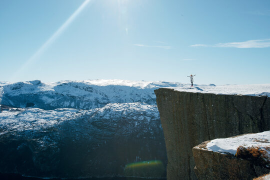 Person standing on snowy cliff edge with arms outstretched, overlooking vast mountain landscape under clear blue sky. Preikestolen, Lysefjord in Norway - Powered by Adobe