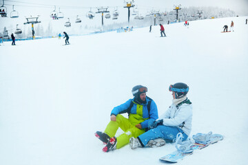 Two friends sit on the snow discussing their ski day at a popular winter resort under cloudy skies...