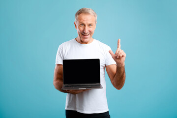 Mature man stands against a blue background, holding a laptop with a black screen. He is smiling and pointing upwards, indicating a sense of excitement and opportunity.