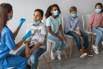 A medical worker performs a PCR test on an African American boy in a clinic. Diverse kids wait their turn while wearing masks in a clean and modern environment.