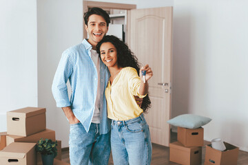Young couple stands smiling in a newly furnished room filled with moving boxes. They hold keys in...