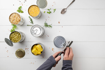 Hands using a manual can opener.  Variety of canned foods on a white table with herbs and spices scattered around.
