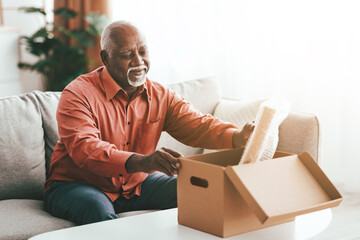 An elderly man with gray hair smiles as he opens a cardboard box on a coffee table in his cozy living room. Sunlight fills the space, creating a warm atmosphere.