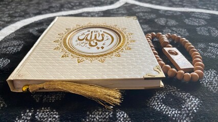 Close-up of a white Quran with gold details, placed on a black and white patterned prayer rug. Beside it is a wooden prayer bead.