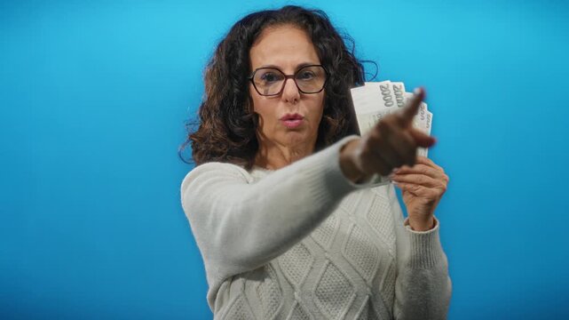 Middle-aged woman holding czech koruna banknotes, pointing emphatically against an isolated blue background, suggesting a financial decision or direction.