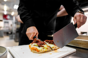 A chef in a black uniform uses two large knives simultaneously to slice a delicious, freshly made mini pizza topped with pulled meat and melted cheese on a white cutting board in professional kitchen