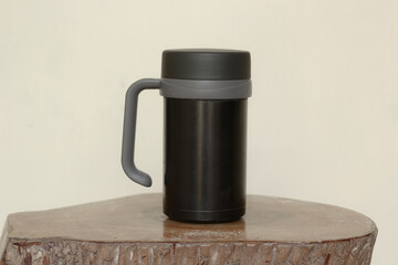 A black travel mug or thermos with a grey handle, standing on a rustic wooden log slice against a plain wall.