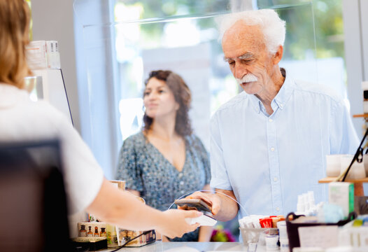 Senior man buying medicine from female pharmacist in pharmacy using smart phone for paying.